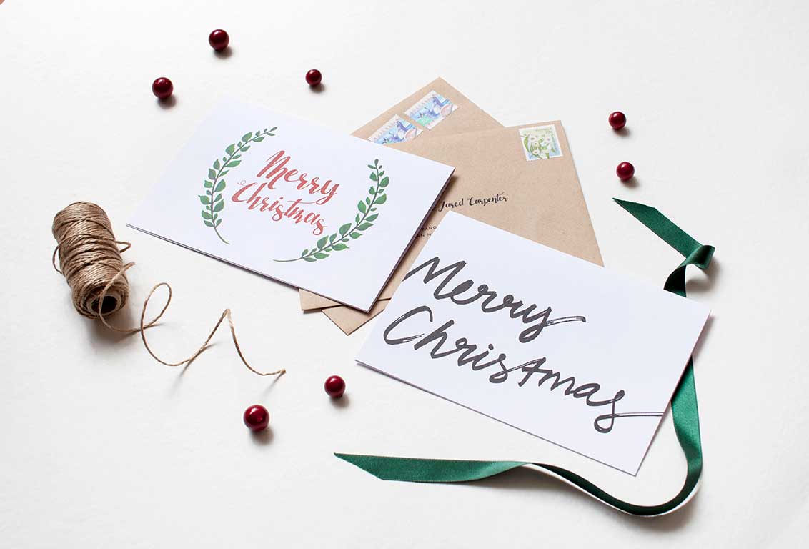 Two illustrated Christmas cards with botanical wreaths and 'Merry Christmas' script, hand addressed kraft envelopes, red baubles, and ribbon.