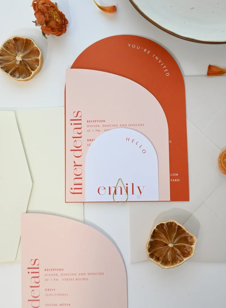 A warm lifestyle photo of a couple reviewing wedding stationery designs together at a table, with a laptop open and stationery samples spread out around them wedding stationery