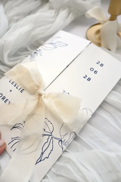 Beautifully styled bridal shower invitation with wavy die-cut edges and pastel pink florals, US address detail visible on card face, professional flatlay on white surface with fresh flowers and ribbon, horizontal composition wedding stationery