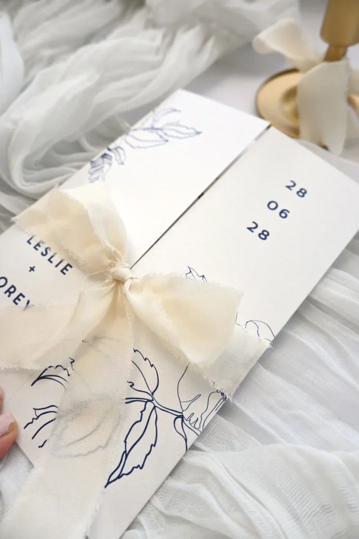 Beautifully styled bridal shower invitation with wavy die-cut edges and pastel pink florals, US address detail visible on card face, professional flatlay on white surface with fresh flowers and ribbon, horizontal composition wedding stationery