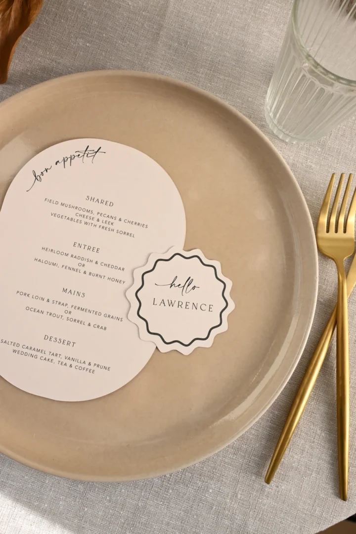 Beautifully styled oval menu card and scalloped-edge place card displayed on a neutral-toned plate at an elegant table setting, minimal composition with soft natural light wedding stationery