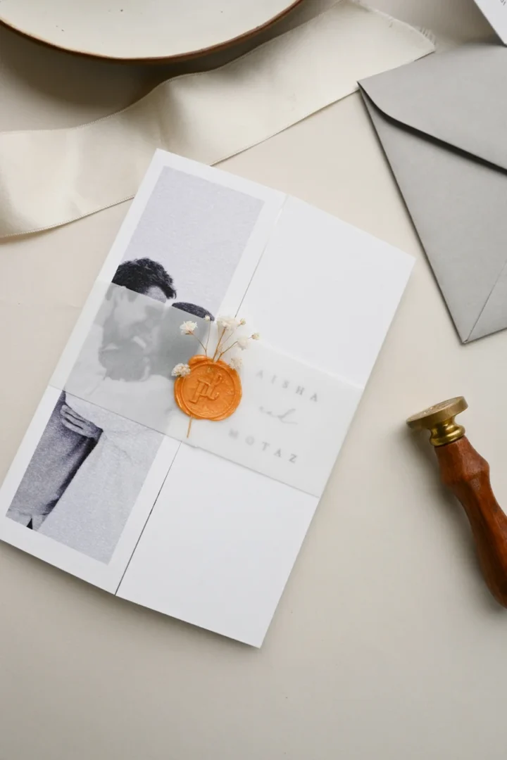 Bridal shower invitation with bold wavy border design in warm red-orange tones, paired with a matching orange envelope and ranunculus flower prop, US address clearly visible, styled flatlay on neutral cream surface wedding stationery