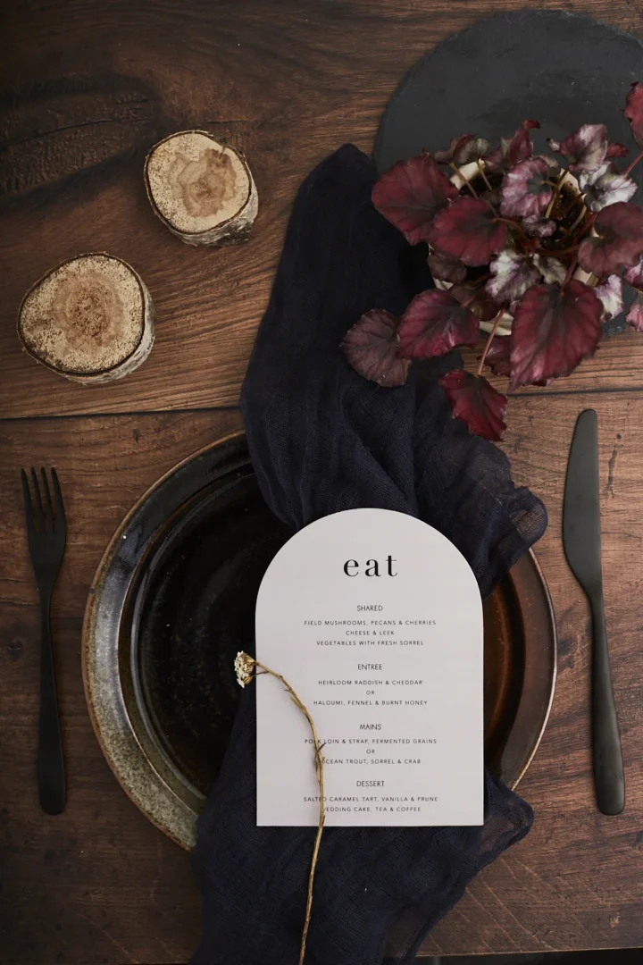 couple sitting together reviewing wedding stationery and planning notes at a rustic wooden table, laptop open with a playlist visible on screen wedding stationery