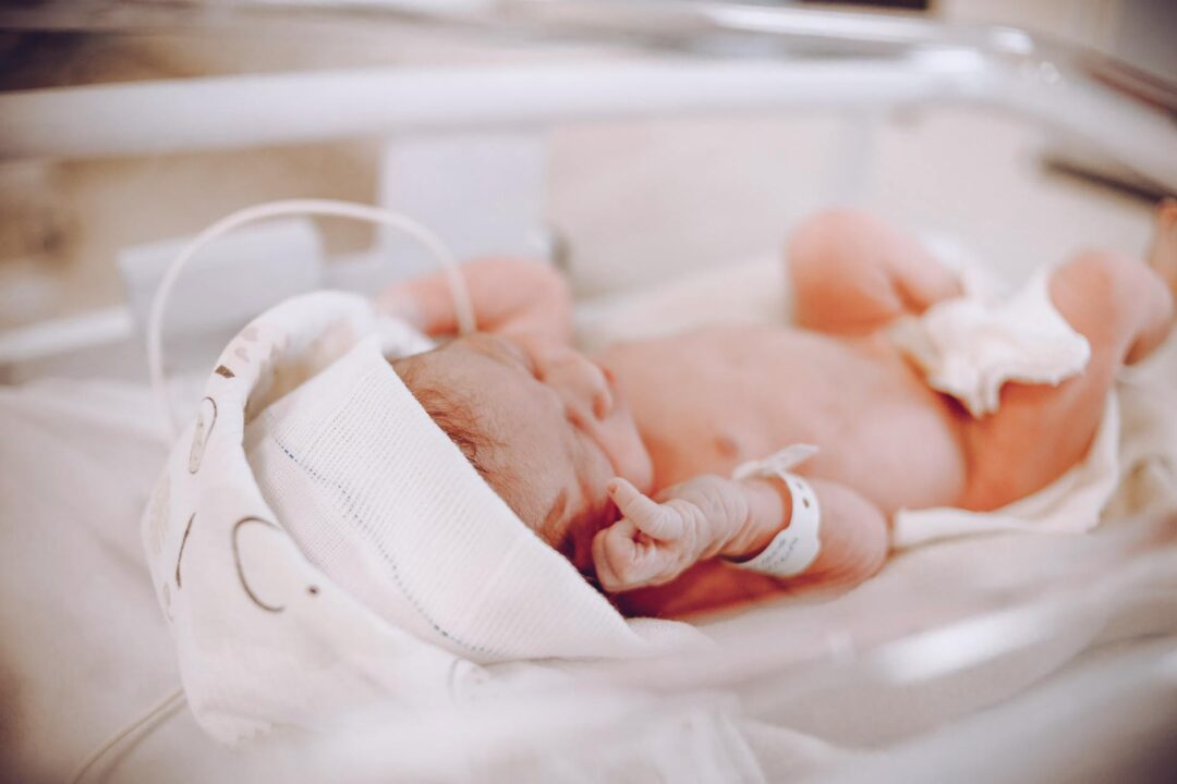 NICU isolette with parent holding hand of premature baby, evoking the timing flexibility for premature births