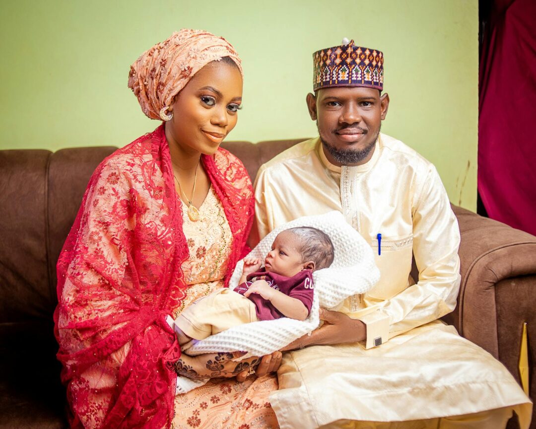 family of three - mother, father, and newborn - in a relaxed first portrait, with photo birth announcement card printed on cotton stock held in mother's hands, soft blurred background