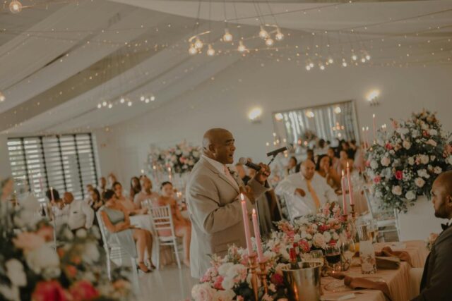best man standing confidently at a wedding reception microphone, guests smiling, warm candlelit ballroom background
