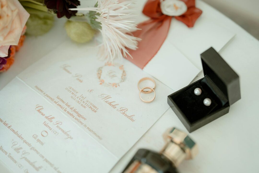 Rose gold wedding bands and pearl earrings sit on an invitation with coral florals and pink ribbon details.