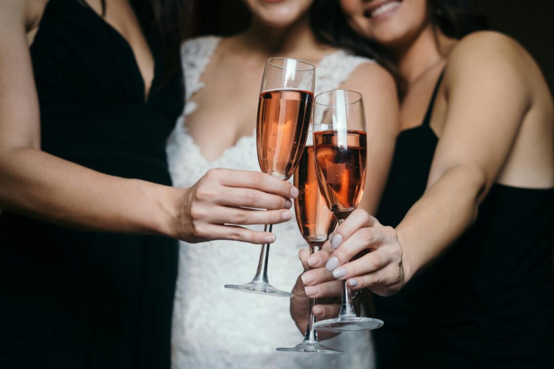Three bridesmaids in blush dresses toasting with champagne flutes at a reception table, natural candlelight in background