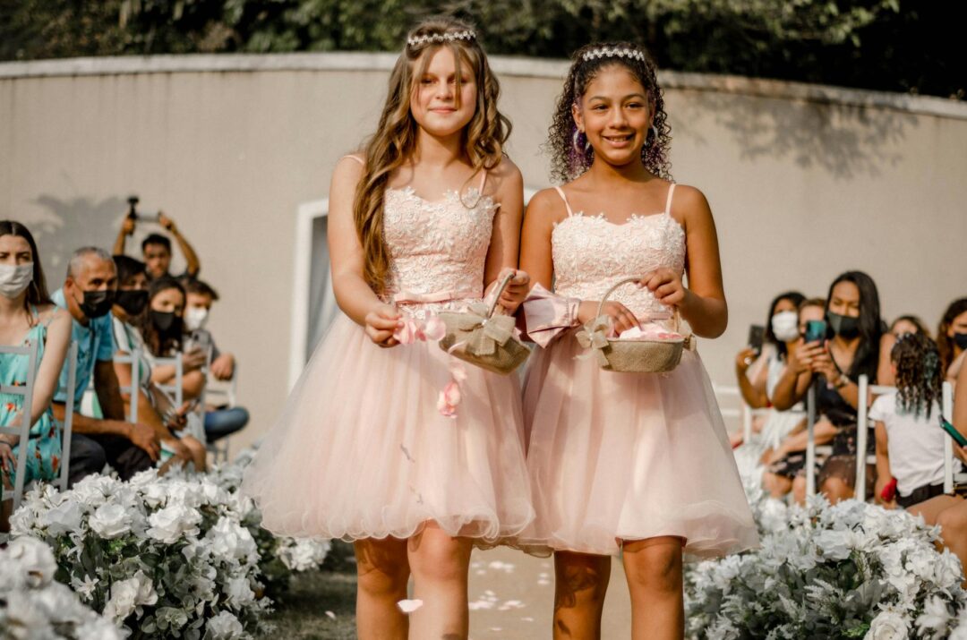 Two young junior bridesmaids in blush dresses walking down a ceremony aisle, carrying small floral pomanders, smiling