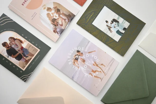 Flat lay of five Christmas cards with family photos in several designs and colors, surrounded by envelopes and fabric swatches on a white background.