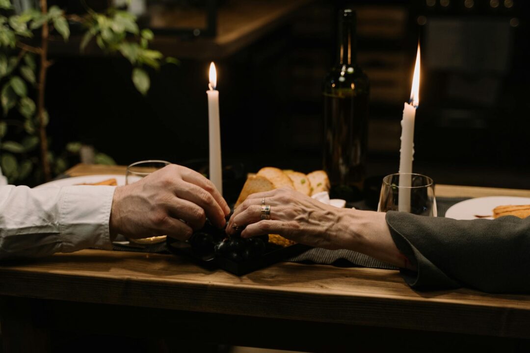 Couple holding hands across a small candlelit table with bread, wine, and grapes, intimate romantic dinner setting that captures the supper-club spirit of a micro wedding. Photo: cottonbro studio / Pexels