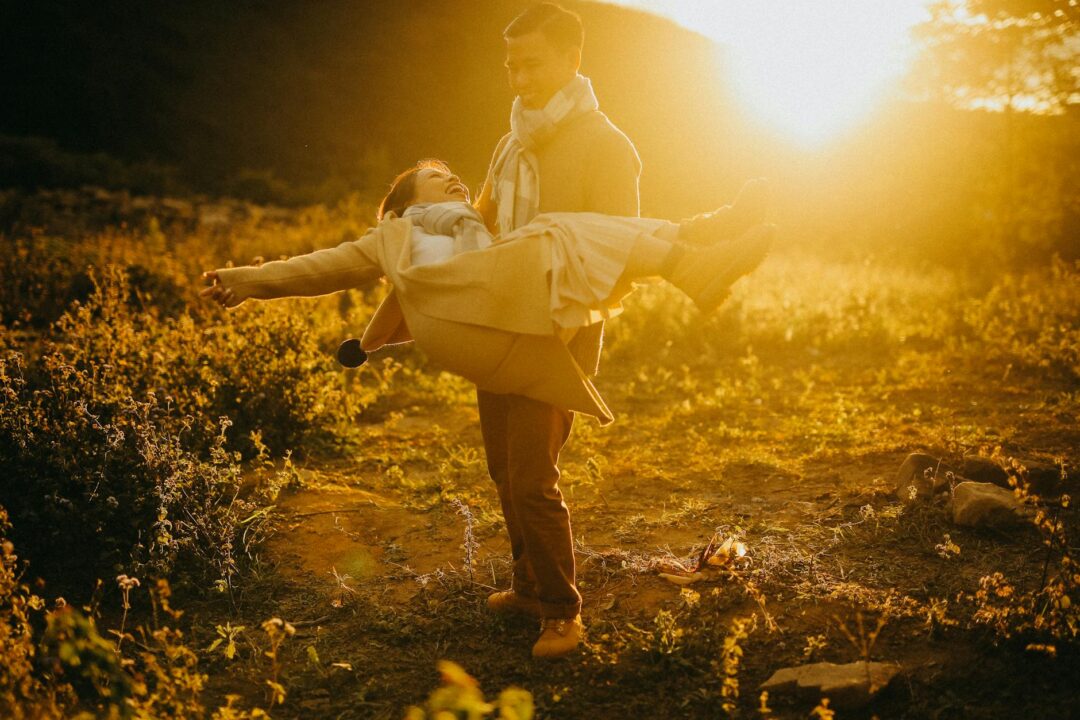 engaged couple laughing and embracing in a sun-dappled meadow at golden hour, warm amber backlight, genuine emotion, wide open field with soft bokeh background