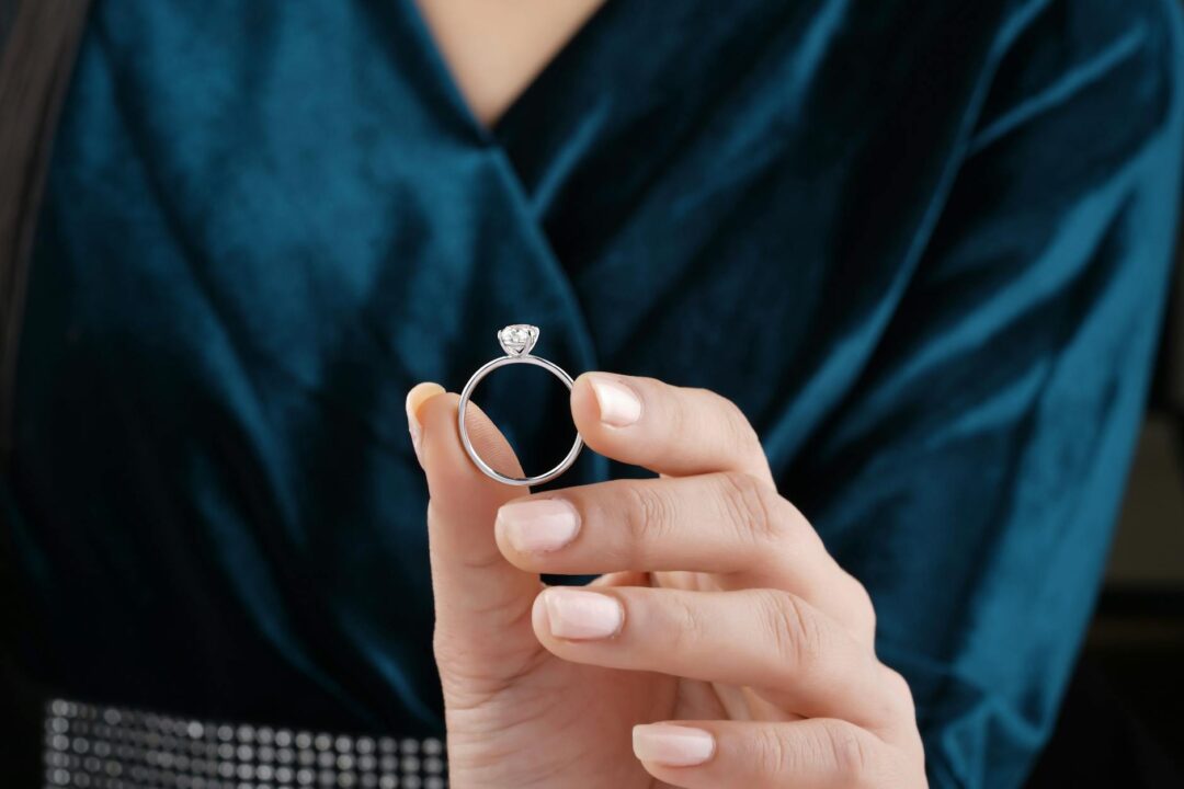 sparkling round brilliant solitaire diamond engagement ring held between fingertips against soft white background, showing facets and light reflection in detail
