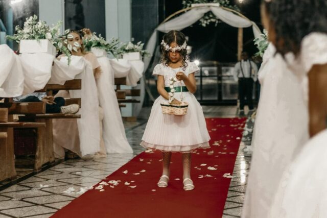 flower girl in a white tulle ball gown walking down a flower-petal-scattered aisle at an outdoor garden wedding, holding a small wicker bask