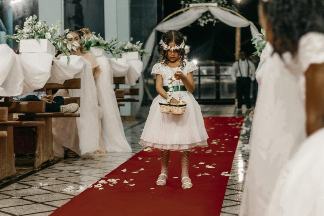 flower girl in a white tulle ball gown walking down a flower-petal-scattered aisle at an outdoor garden wedding, holding a small wicker bask
