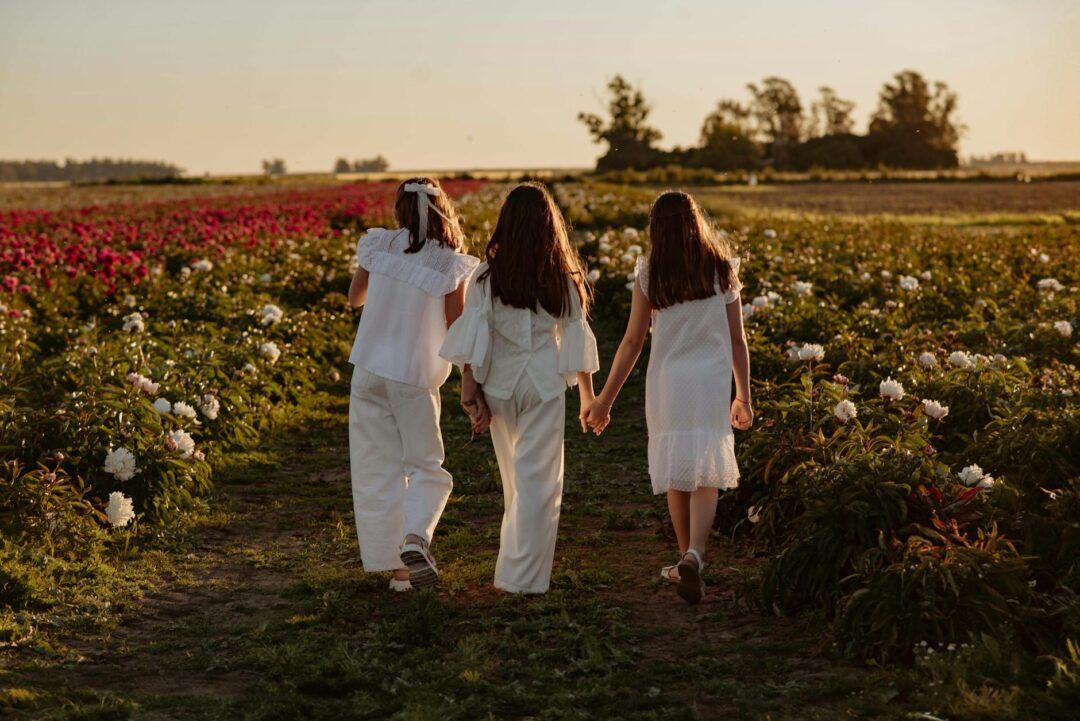 three flower girls of different ages standing together in coordinating white and cream dresses at a garden wedding, smiling naturally, edito