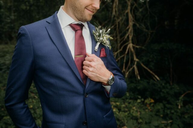 groom in a navy three-piece suit standing at an outdoor wedding altar surrounded by greenery, boutonniere pinned to lapel, confident relaxed