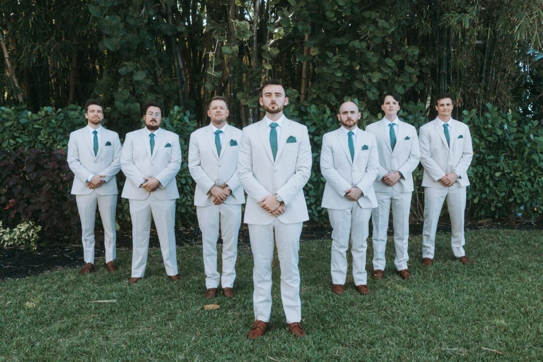 groom in sage green suit standing outdoors in garden setting, holding ceremony program, soft natural light filtering through trees behind hi