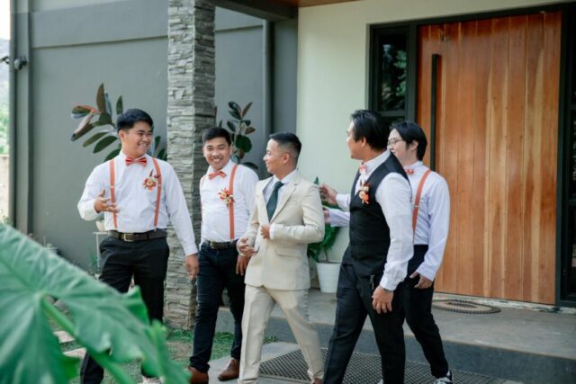 groom and groomsmen laughing together outdoors at a sunlit wedding venue, dressed in matching suits, one holding a whiskey glass in a toast