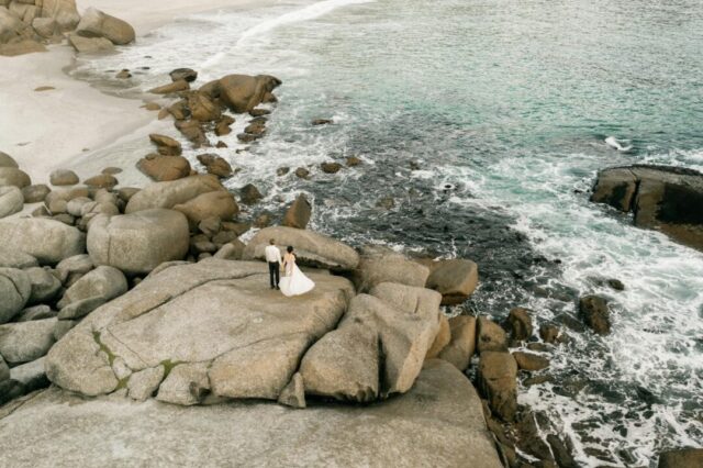 Newlywed couple standing on a sun-drenched cliffside terrace overlooking the Amalfi Coast, champagne flutes raised, turquoise Mediterranean sea stretching behind them