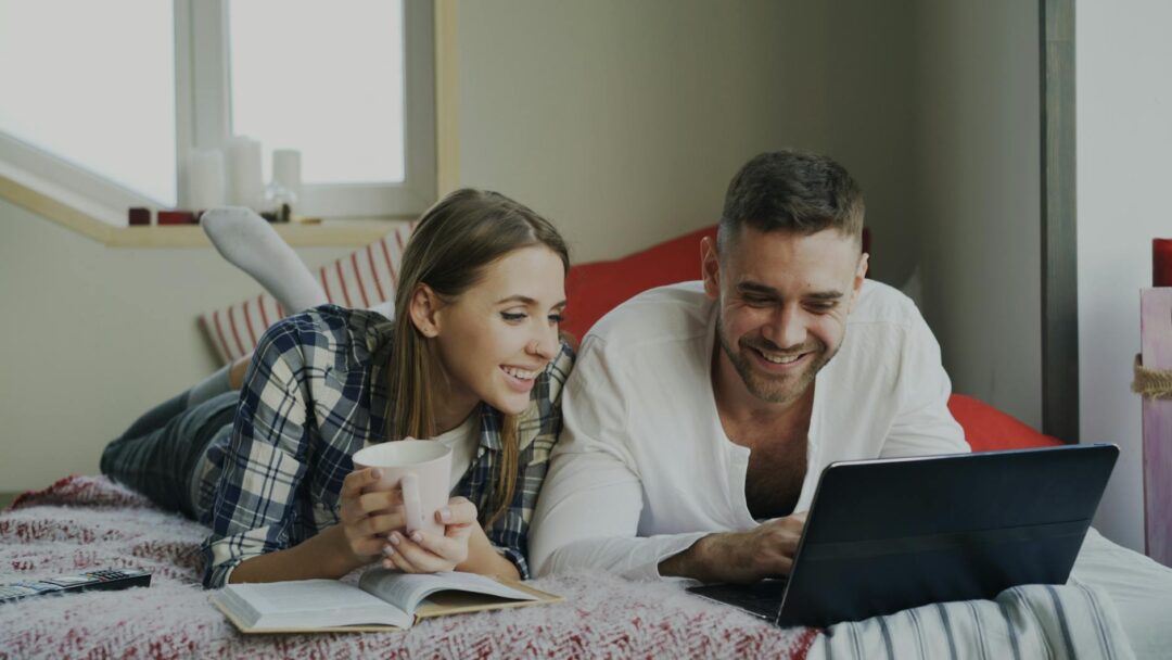 couple browsing hotel booking website on laptop with travel brochures and a honeymoon planning notebook beside them