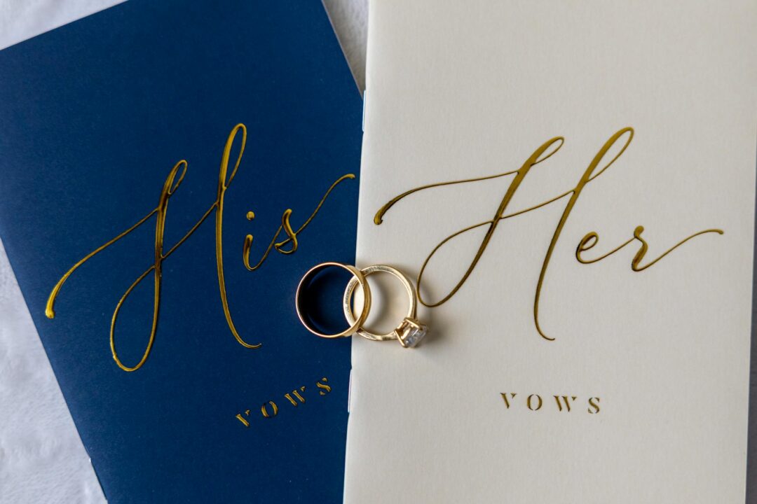 An overhead flatlay of a wedding ceremony script binder open to a vows page, with a pen and small succulent beside it on a white linen surfa