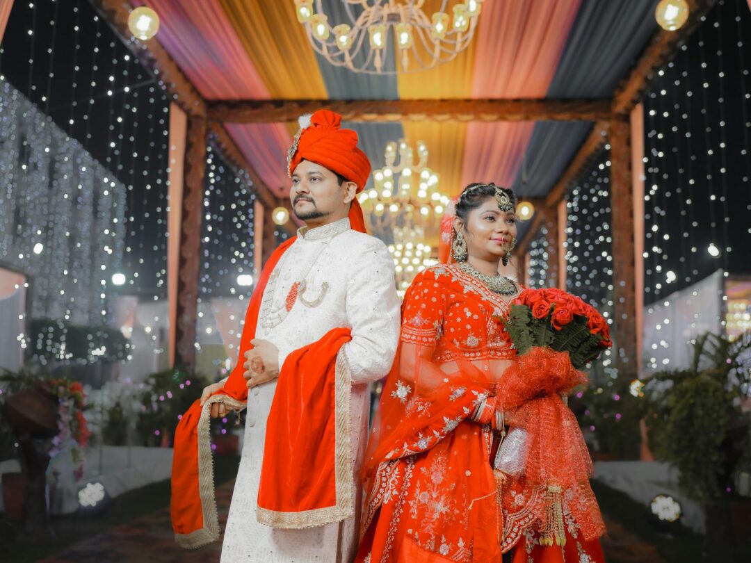 Bride and groom standing under a floral mandap canopy decorated with marigolds and roses, both dressed in red and gold, during the exchange