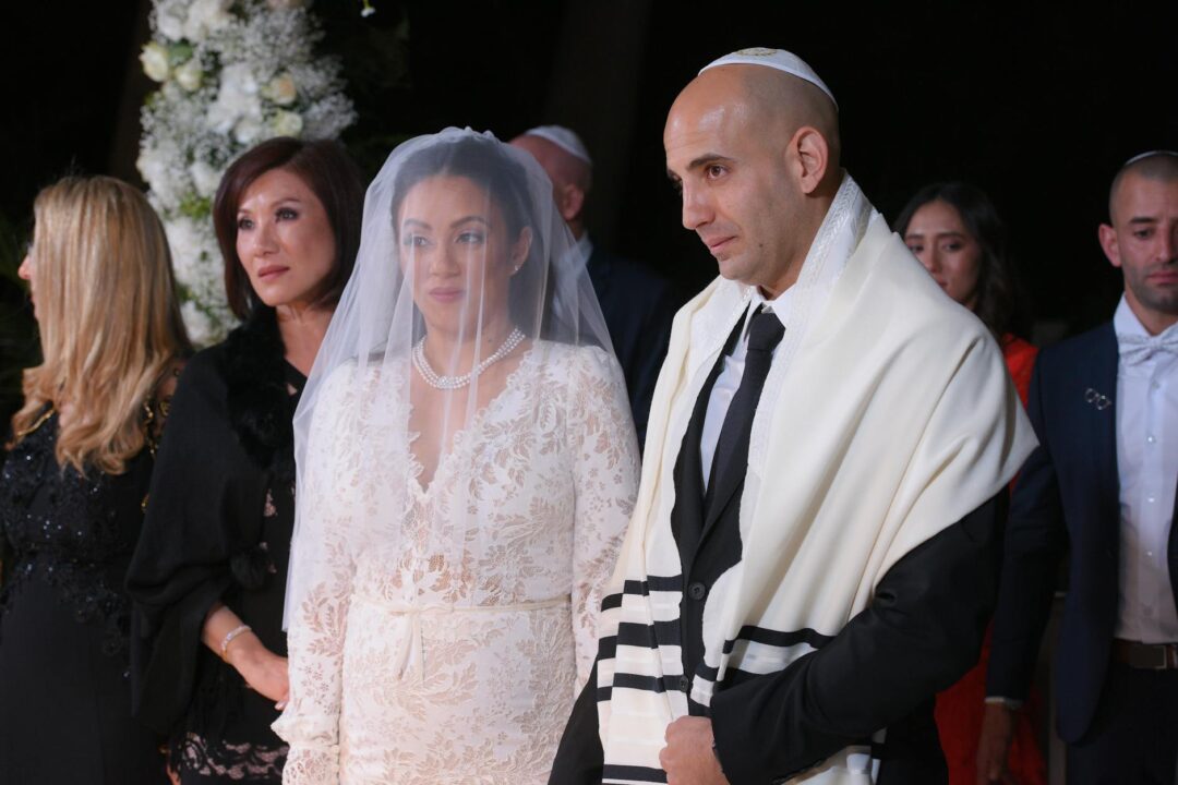 Jewish wedding ceremony beneath a floral chuppah canopy decorated with white roses and greenery, couple standing together under soft natural