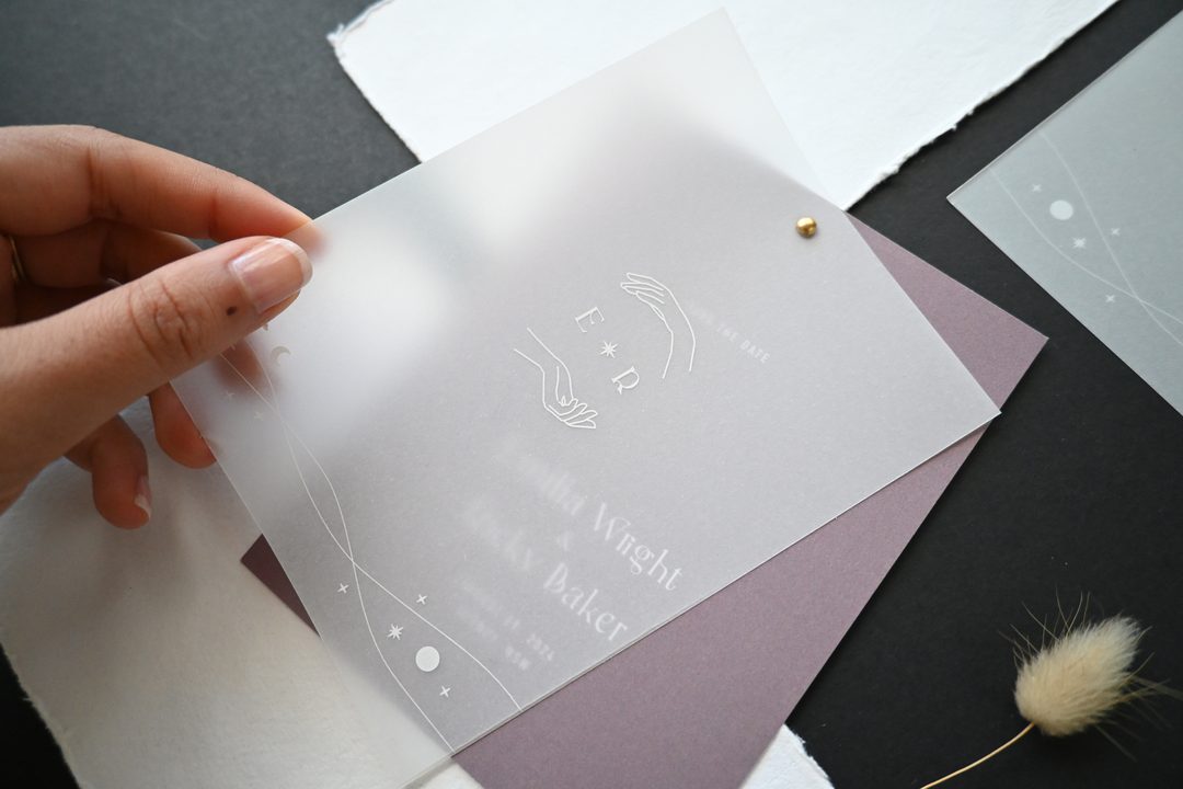 Paperlust vellum save the date card with white foil celestial line art held up to show layered translucent overlay over a dusty mauve base card