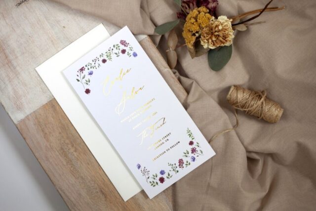Paperlust Amaranth slim DL wedding invitation -- white card with watercolour wildflower border in burgundy florals and sage green botanical, gold foil calligraphy script, styled on a wood board with cream envelope and dried yarrow