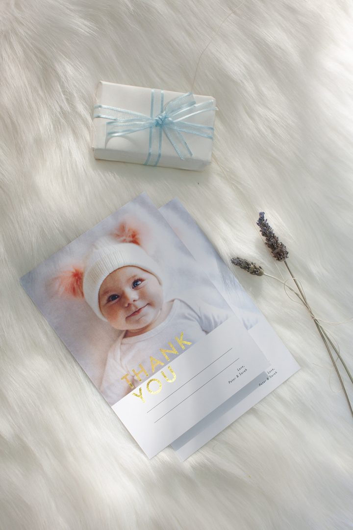 Paperlust portrait-format birth announcement card with baby photo and gold foil script, styled on a fluffy white rug alongside a ribbon-tied gift