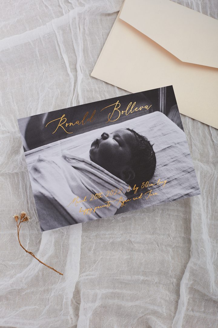Paperlust landscape birth announcement card with full-bleed black and white newborn photo and gold foil script typography, paired with a cream envelope
