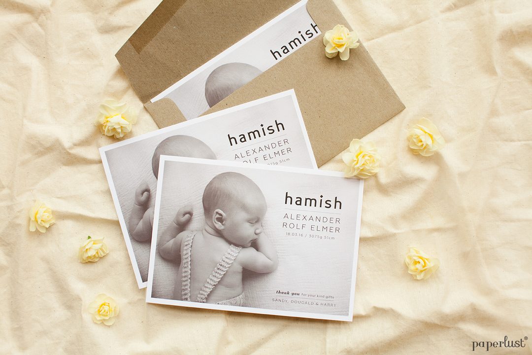 Paperlust landscape birth announcement card with black-and-white newborn photo paired with a kraft paper envelope, on a neutral flatlay surface