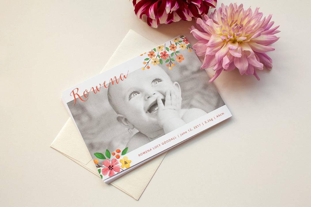 Paperlust landscape birth announcement card with watercolour floral border, coral script typography, and a black-and-white baby photo, resting on a cream envelope