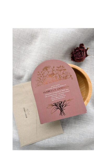 Paperlust arch-shaped dusty rose bridal shower invitation with rose gold foil botanical illustrations on a kraft envelope in a wooden bowl