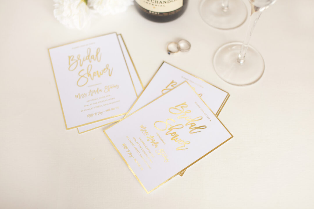 Paperlust three square white bridal shower invitations with gold foil script typography fanned on cream surface alongside champagne