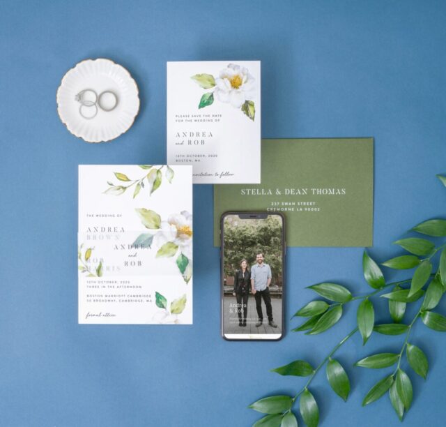 Paperlust hand-illustrated botanical wedding invitation suite -- white cards with watercolor white magnolia blooms and green leaves, sage envelope, mobile mockup, styled on muted blue background with eucalyptus