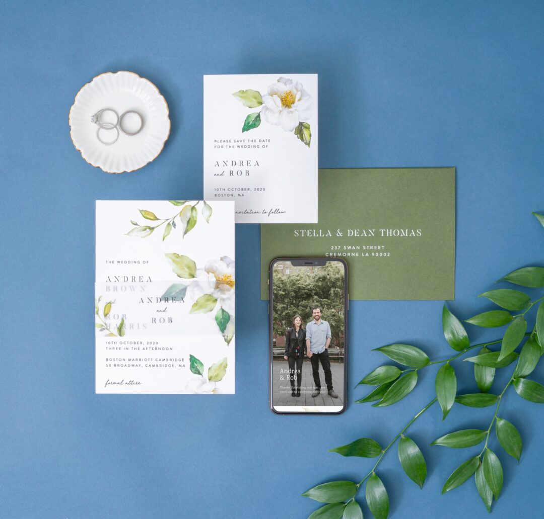 Paperlust hand-illustrated botanical wedding invitation suite -- white cards with watercolor white magnolia blooms and green leaves, sage envelope, mobile mockup, styled on muted blue background with eucalyptus
