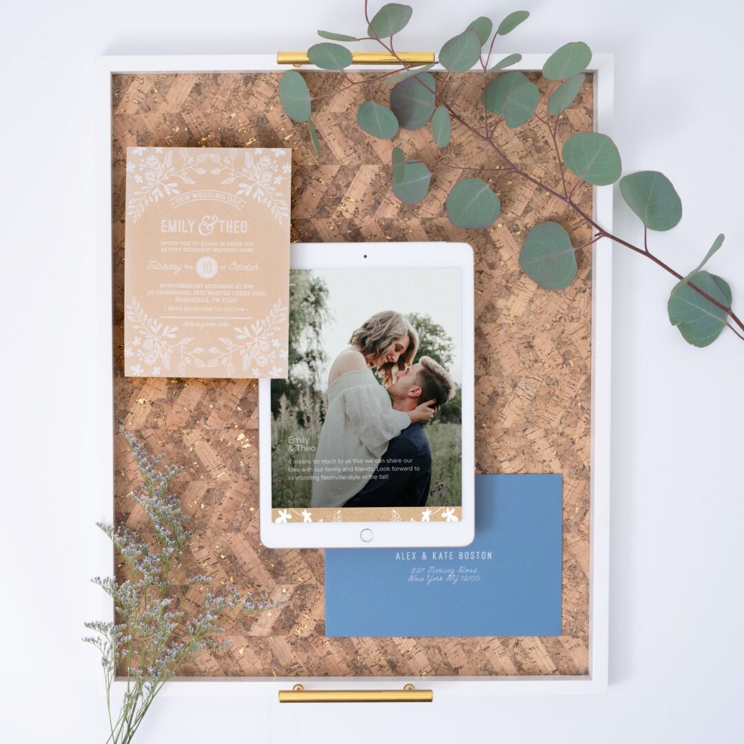Paperlust kraft paper wedding invitation, recycled kraft cardstock with white botanical illustration and warm cream typography, dusty blue envelope, mobile mockup, eucalyptus styling on cork tray