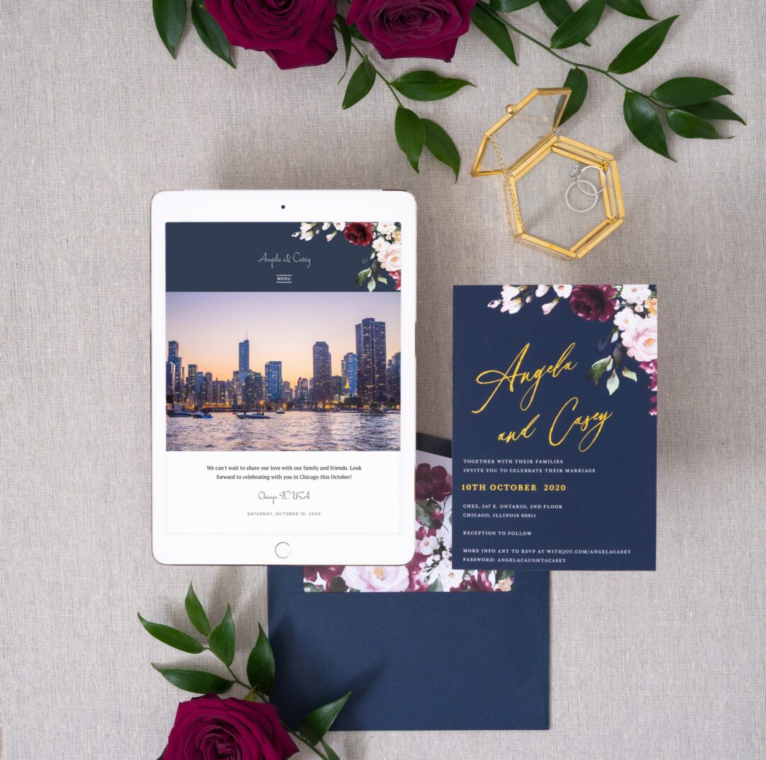 Paperlust La Butte navy and gold wedding invitation suite, navy blue invitation card with gold foil script names and burgundy and blush watercolor floral accents, navy envelope, styled flatlay with burgundy roses and gold ring box