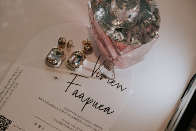 Paperlust vellum arch wedding invitation -- frosted translucent arch card with elegant black script "Vivien + Faapua" calligraphy, styled with crystal earrings and Versace perfume