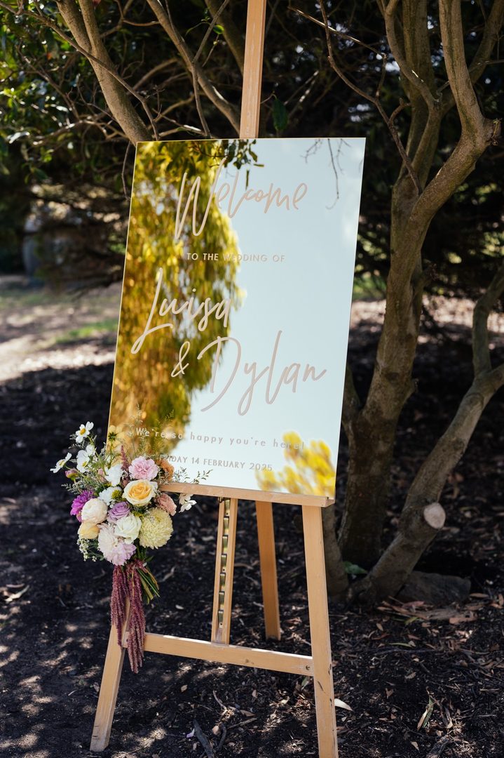 Paperlust welcome sign, Gold mirror acrylic welcome sign for Luisa & Dylan on wooden easel with colorful floral arrangement. Date reads Friday 14 February 2025 (DD Month YYYY format).