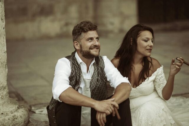 A couple in elegant semi-formal outfits laughing together in a sun-drenched urban alley, the woman in a flowing champagne dress and the man