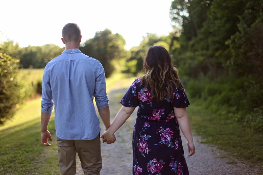 A couple holding hands walking through a lush botanical garden path lined with flowering shrubs, dressed in coordinated neutral tones