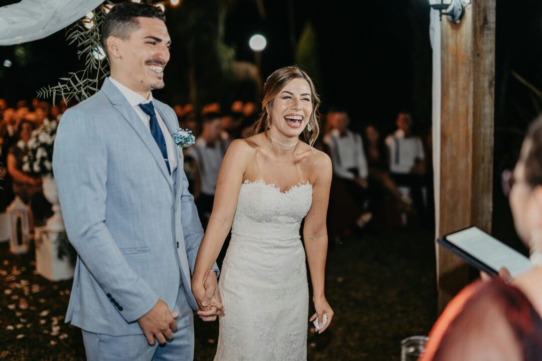 couple laughing joyfully at the altar during vow exchange, guests visibly amused, outdoor garden ceremony, bright natural light, candid reaction shot