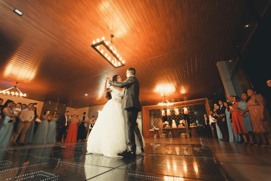bride and groom sharing their first dance on a softly lit ballroom dance floor, fairy lights overhead, guests watching from round tables in the backgr