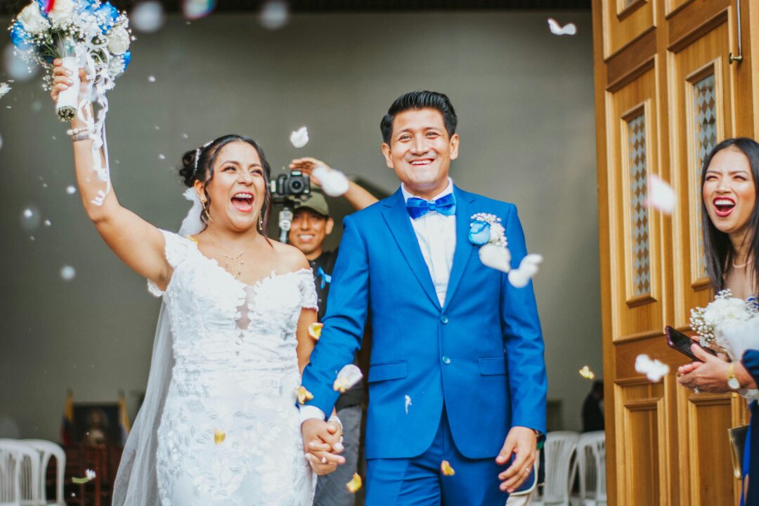 just-married couple running jubilantly down a confetti-filled outdoor aisle, guests cheering and clapping, golden afternoon light