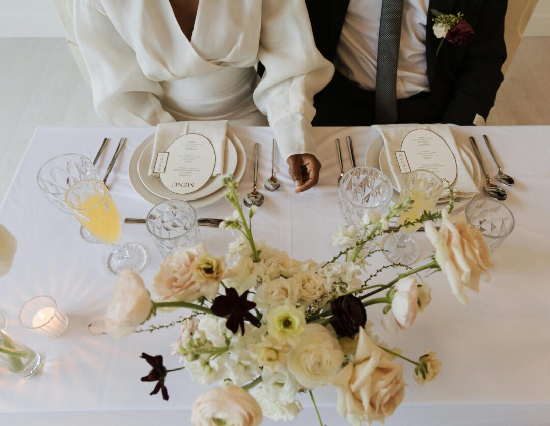overhead flat-lay of an elegant wedding reception dinner table fully set with white linens, floral centerpieces, fine china, and gold cutlery - warm c