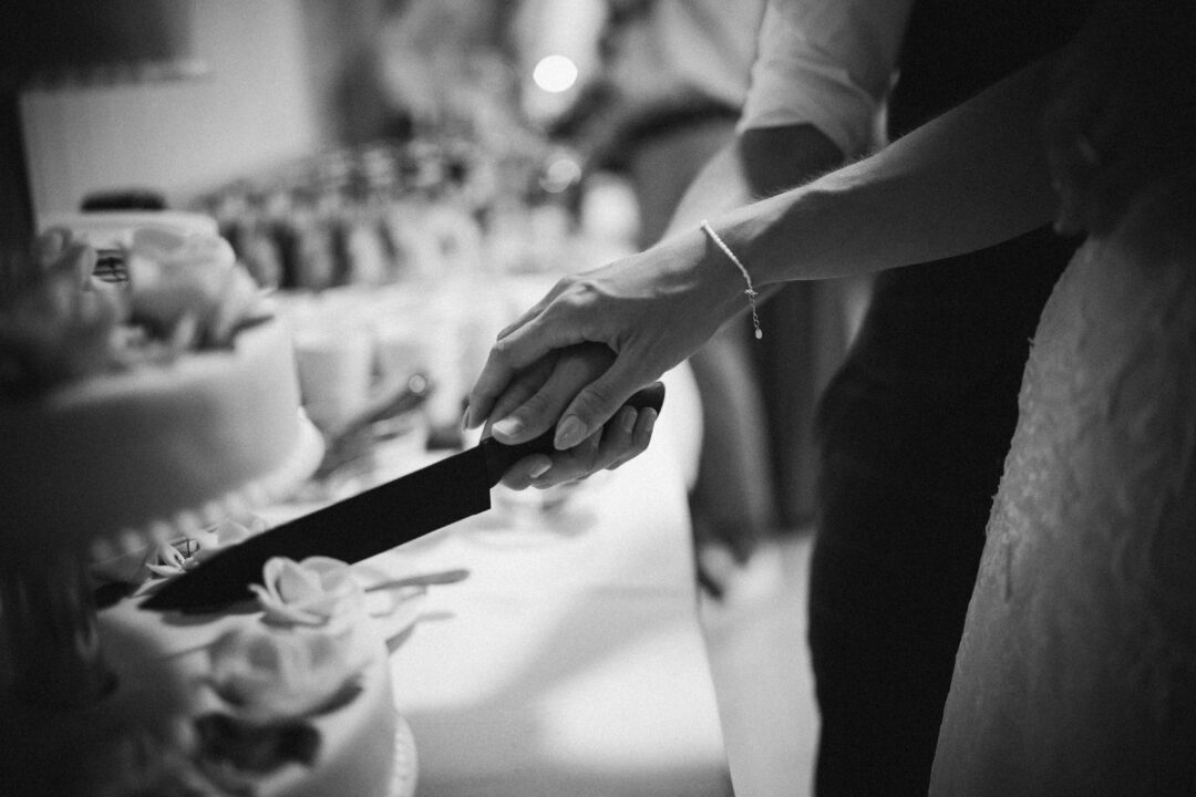 close-up of a bride and groom's hands together cutting a white fondant wedding cake with fresh flowers on top