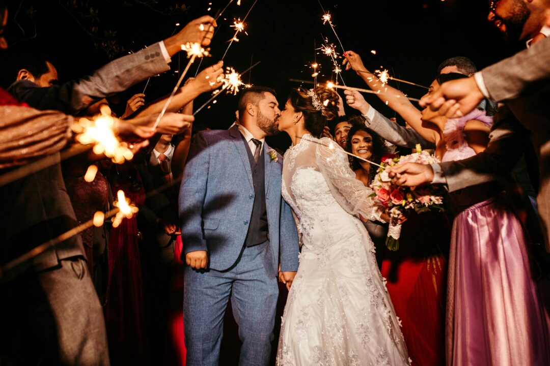 newlywed couple running under a tunnel of sparklers held by guests at dusk, big smiles, bokeh lights in background